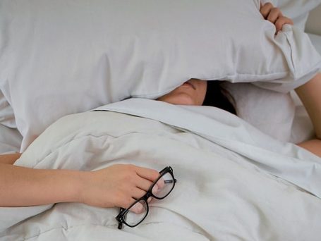 schlafende Frau unter weißer Bettwäsche mit Brille in der Hand