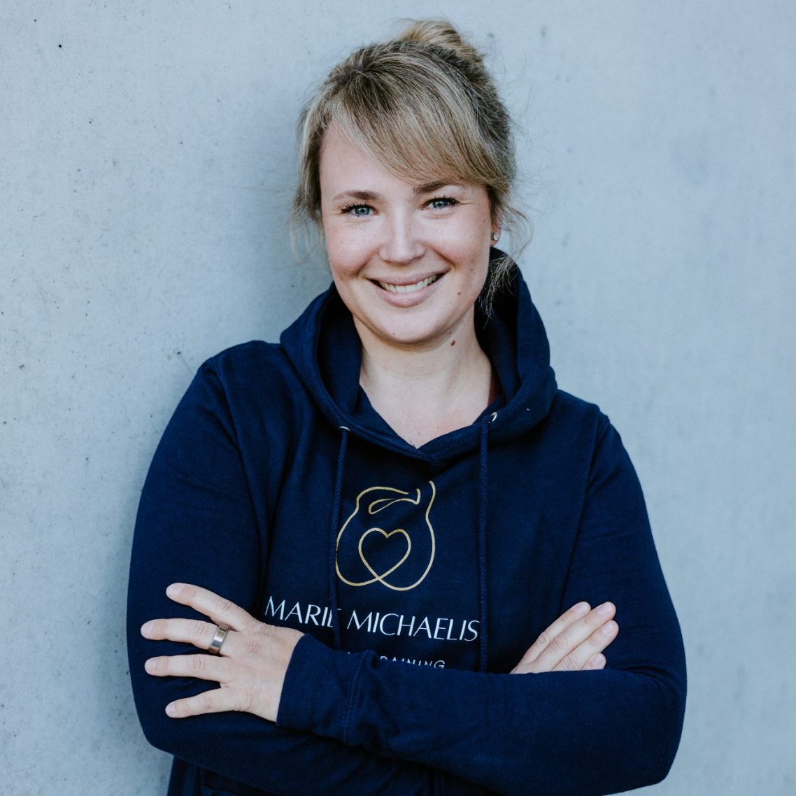 Lächelnde Frau mit blonden Haaren in einem blauen Hoodie, Arme verschränkt vor einer grauen Wand.